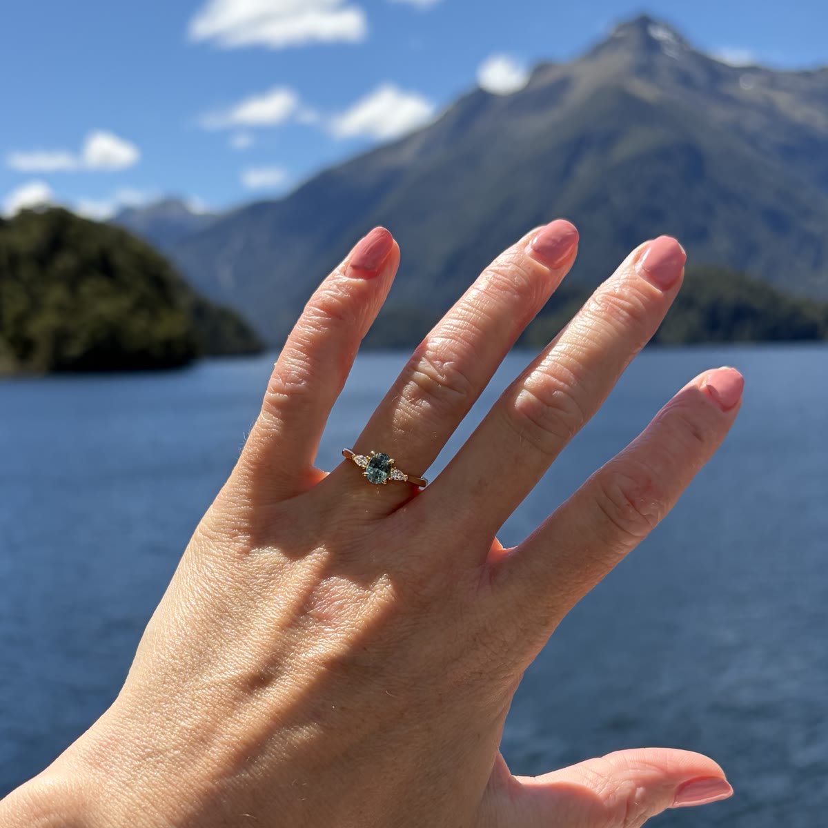 Teal sapphire engagement ring held beside a mountain lake